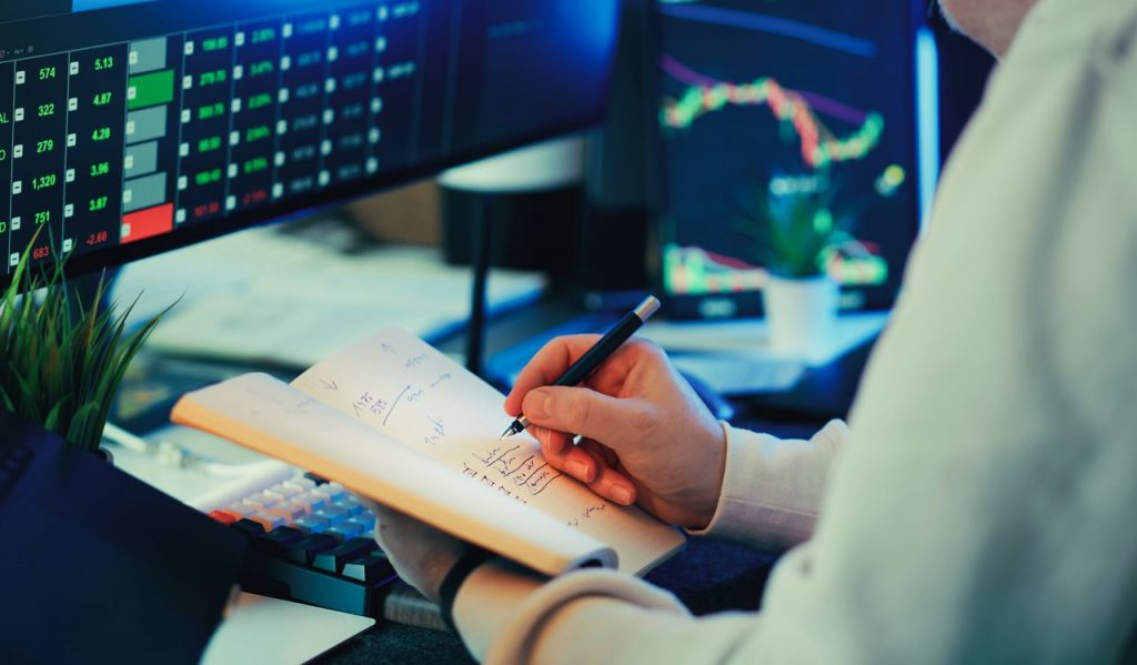Person analyzing financial data on screens, making notes